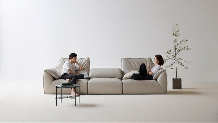 Two people sitting on a modern beige sofa in a minimalistic room with a plant.